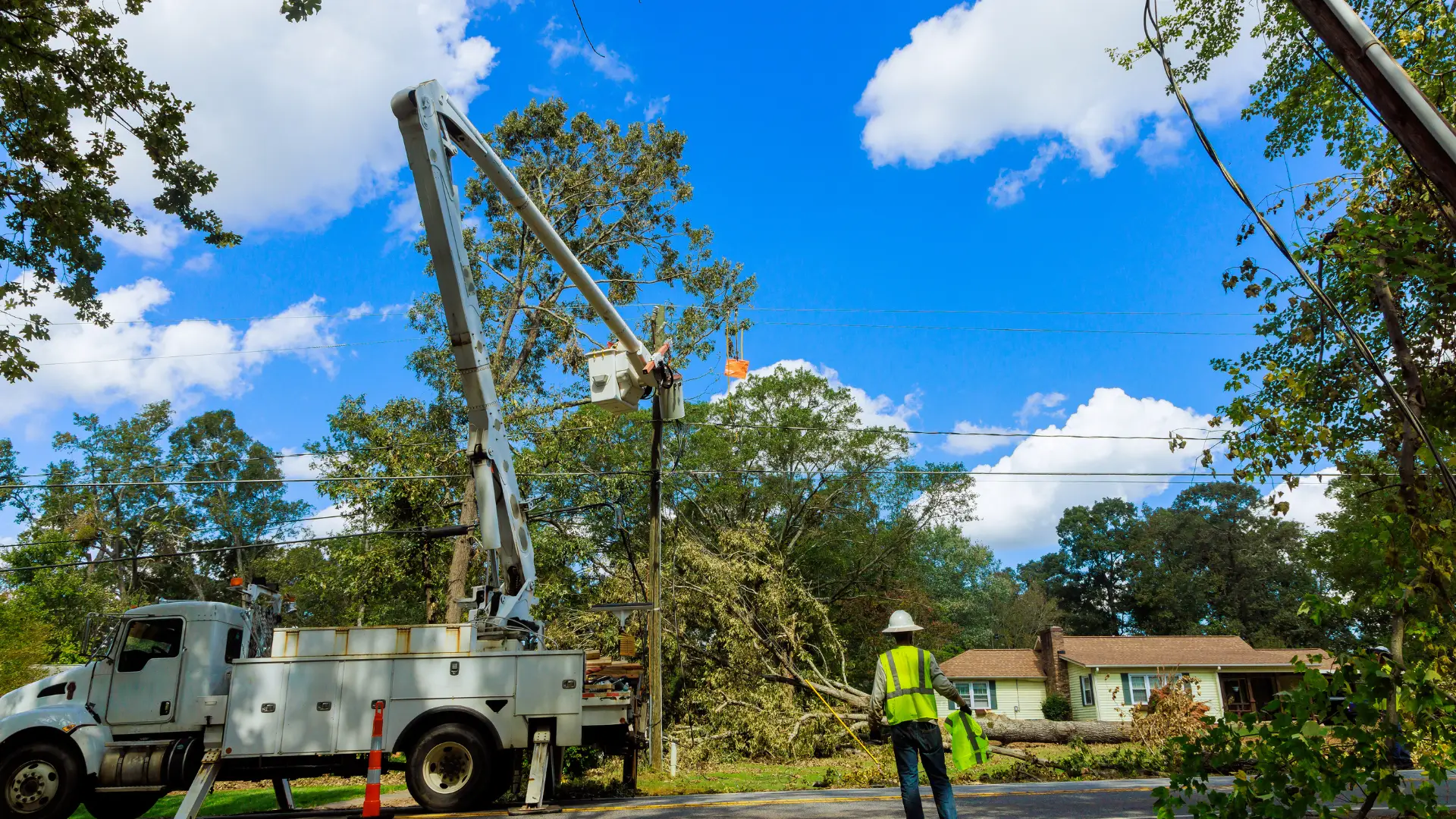 A utility worker in a safety vest and hard hat stands near a bucket truck, overseeing restoration efforts as another worker uses the bucket to repair power lines. A fallen tree lies across the road in this suburban neighborhood.