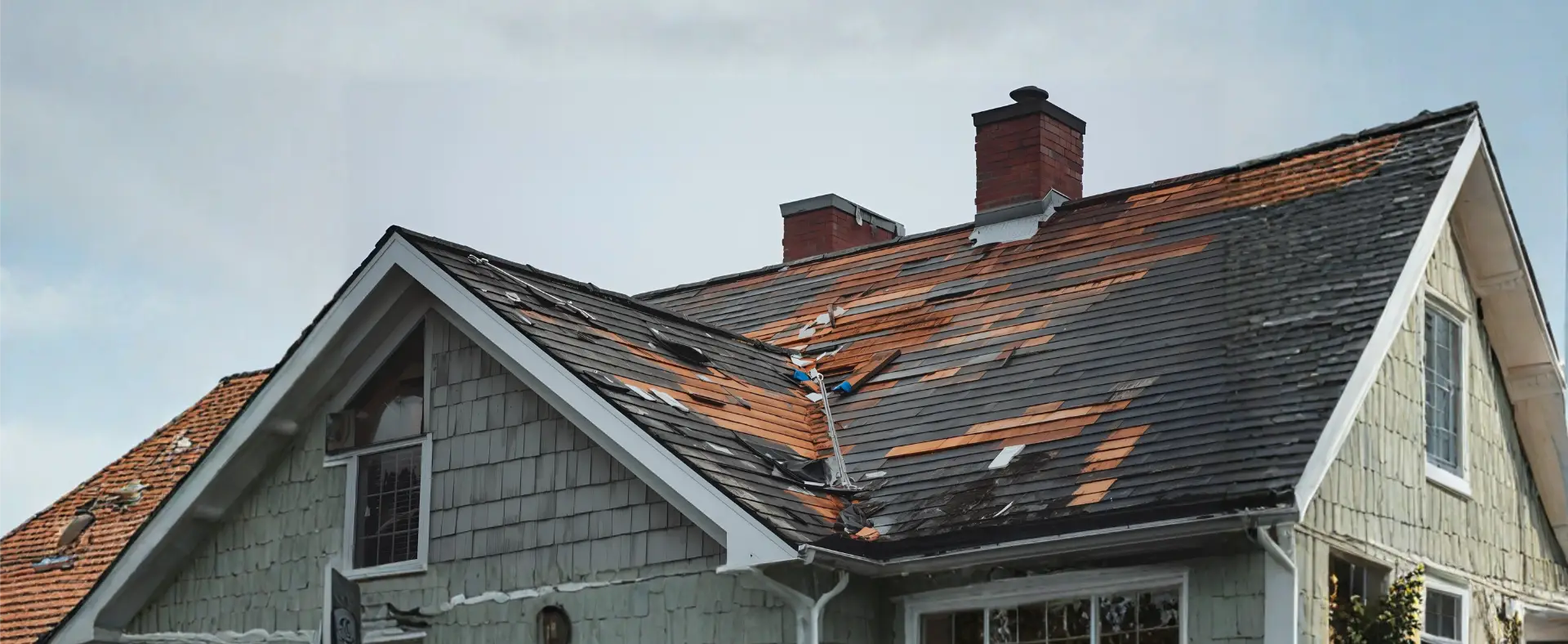 A house with a damaged roof missing many shingles, exposing the underlying materials, shows signs that may require Flood Damage Restoration. Two red brick chimneys are visible beneath the overcast sky.