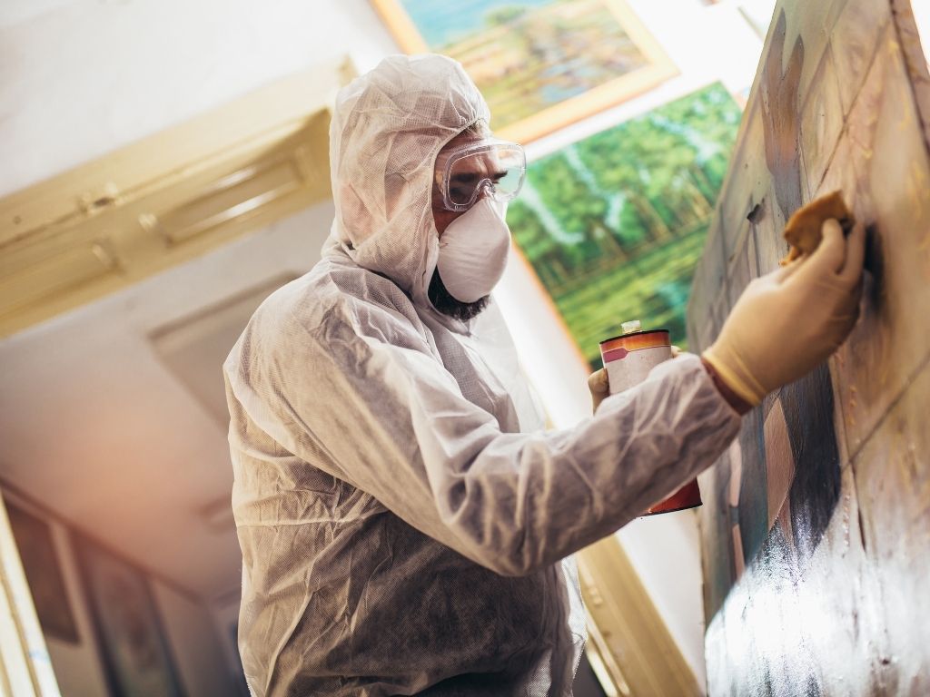 A person in protective coveralls, gloves, goggles, and a mask carefully restores a painting affected by fire damage restoration in an art studio near Marion IL, gently cleaning its surface with framed artworks visible in the background.