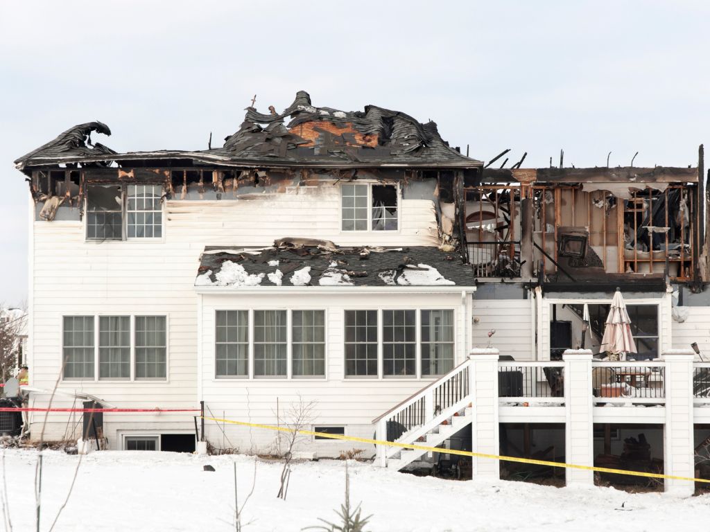 A two-story white house in Marion IL shows severe fire damage to the roof and upper floor, exposing the interior. Snow covers the ground, and caution tape is seen in the foreground—reminding of the importance of Fire Damage Cleanup.